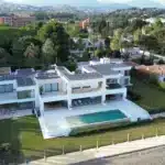 A large modern white villa with multiple balconies and a swimming pool, surrounded by greenery and neighboring houses.