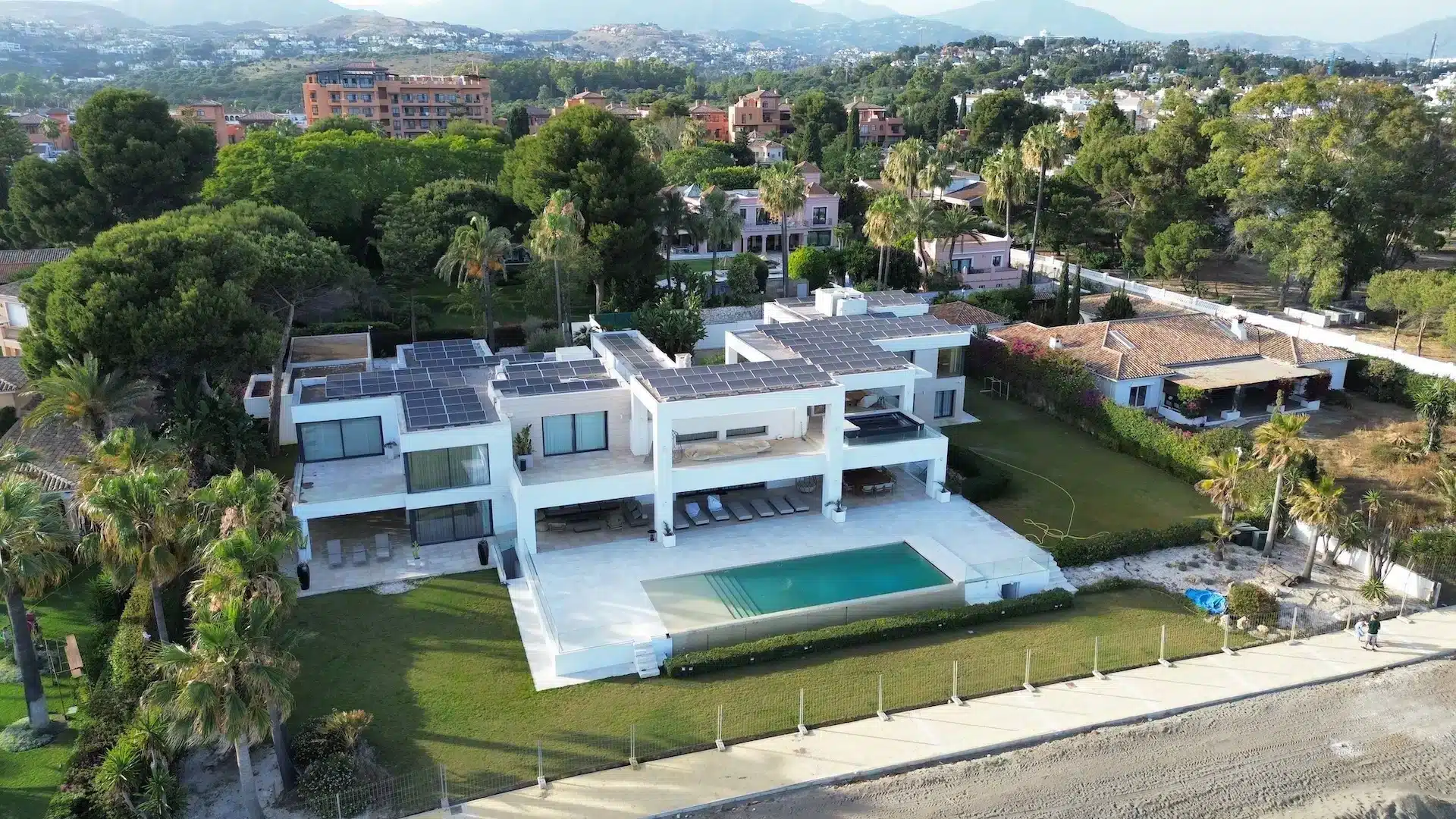 Una gran villa blanca y moderna con múltiples balcones y piscina, rodeada de vegetación y casas vecinas.