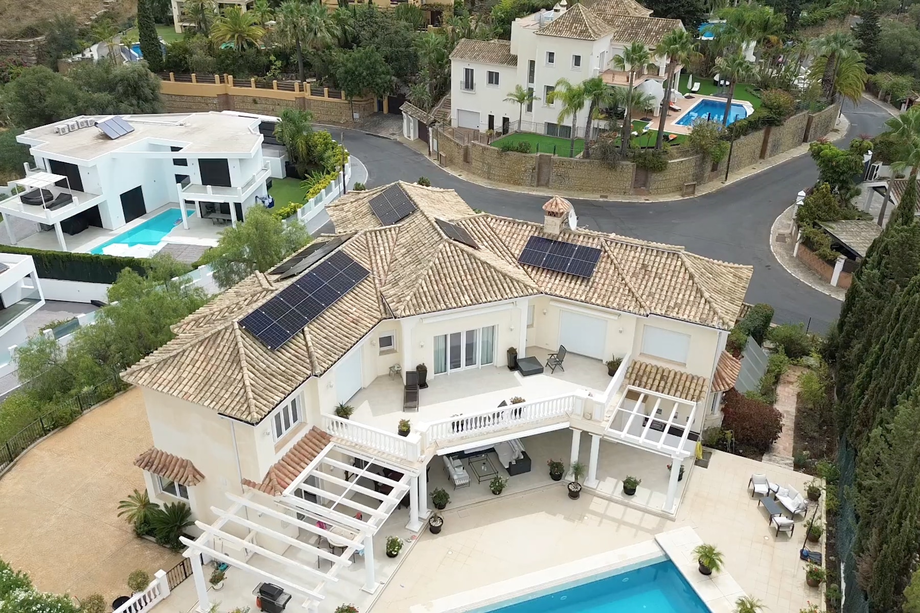 A large white house with solar panels on the roof and a spacious balcony overlooking a pool.