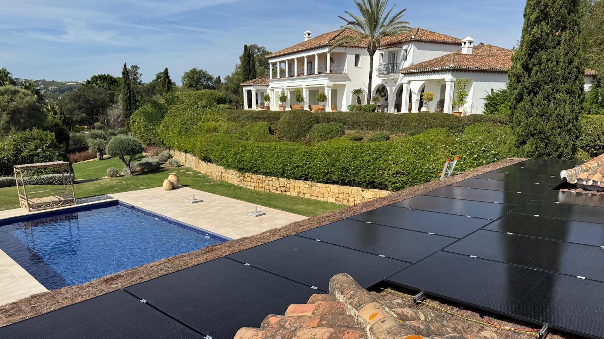 A luxurious white villa with a large balcony overlooking a swimming pool and manicured garden under a clear sky.