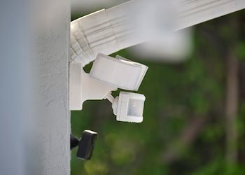 Close-up of a white outdoor security camera mounted on a white wall with greenery in the background.