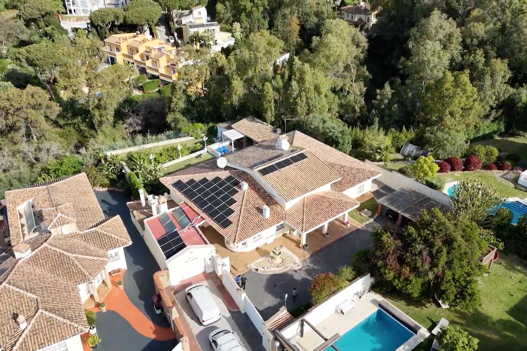 A large house with solar panels on the roof, surrounded by trees and featuring a swimming pool.
