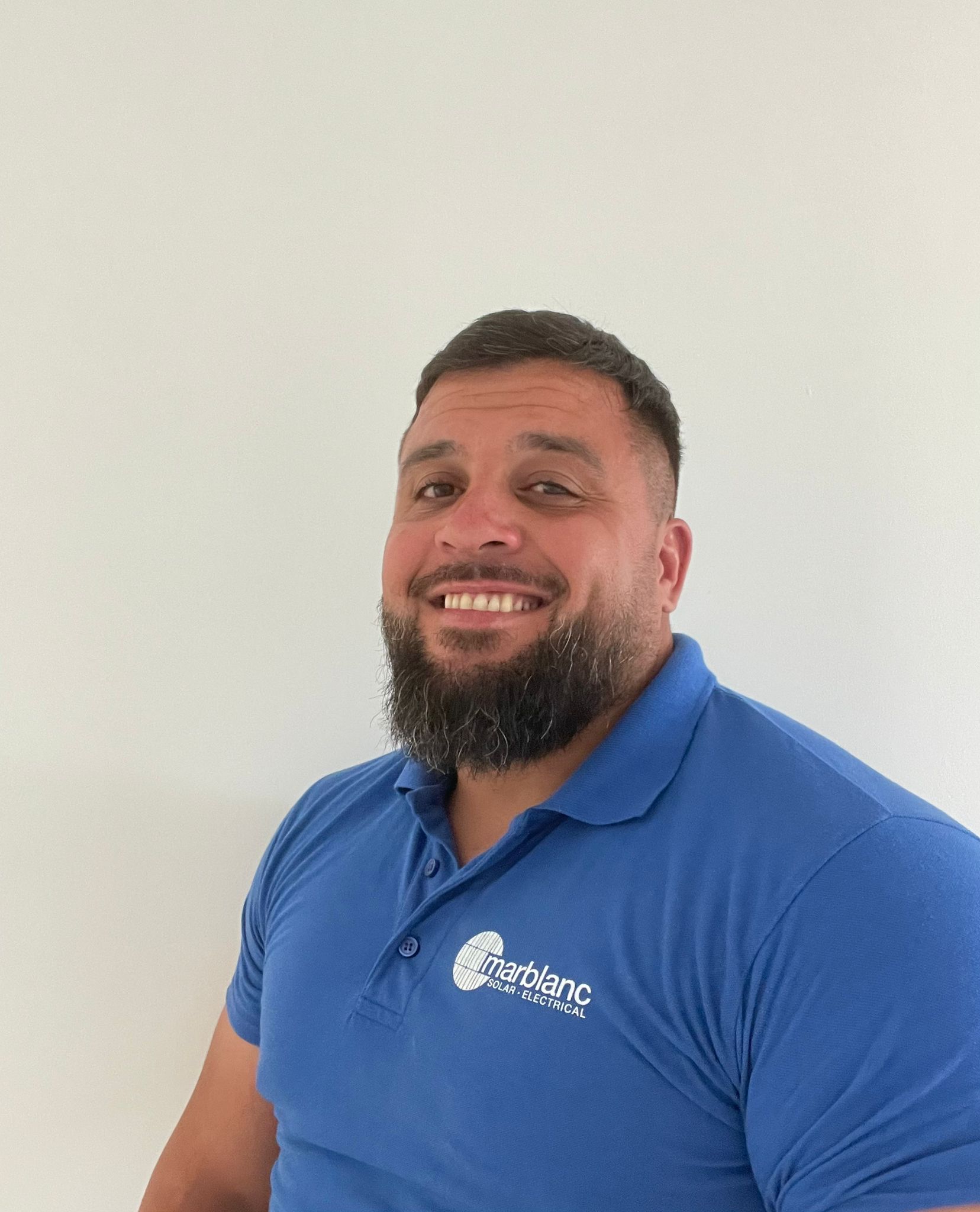 A smiling man with a beard wearing a blue polo shirt with a logo, standing against a plain white background.