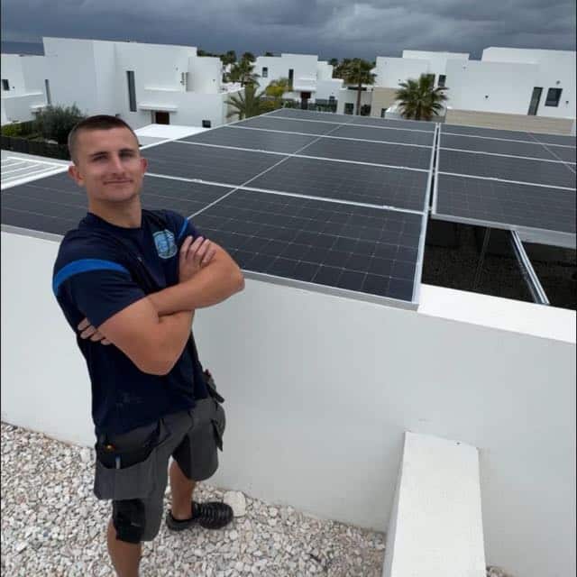 A man stands confidently with arms crossed next to rooftop solar panels under a cloudy sky.