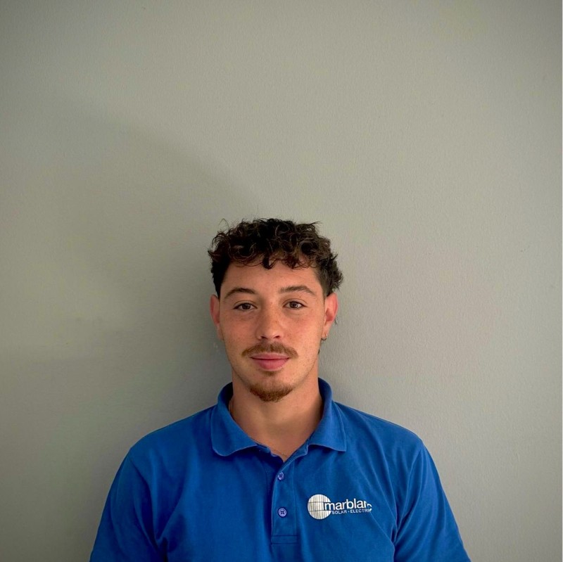 A young man with curly hair wearing a blue polo shirt standing against a plain gray wall.