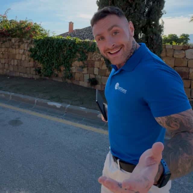 A smiling man in a blue shirt and shorts reaching out towards the camera on a sunny street.