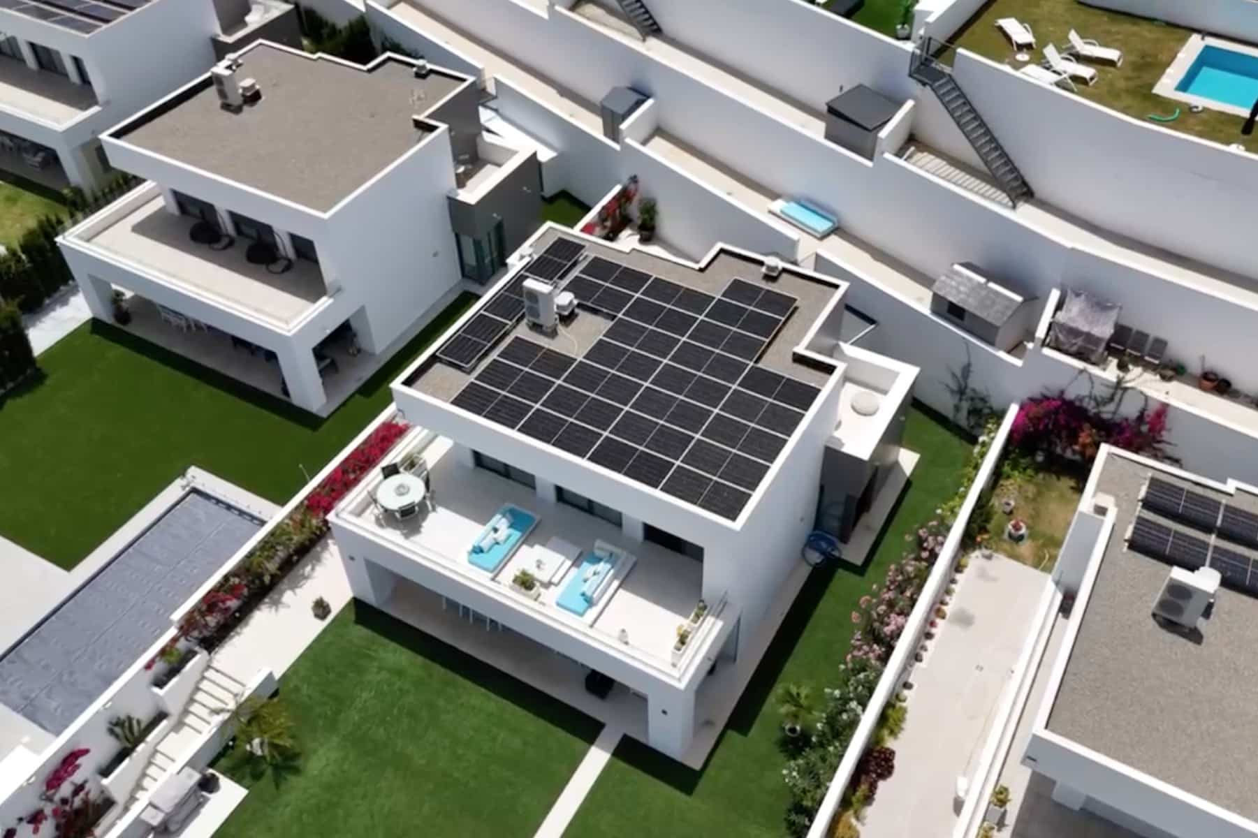 Una moderna casa blanca con paneles solares en el tejado y una piscina en la azotea, rodeada de casas de estilo similar y césped verde.
