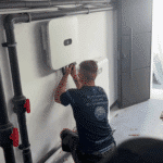 A man crouched down, working on or inspecting a white wall-mounted device with pipes around it in a room.