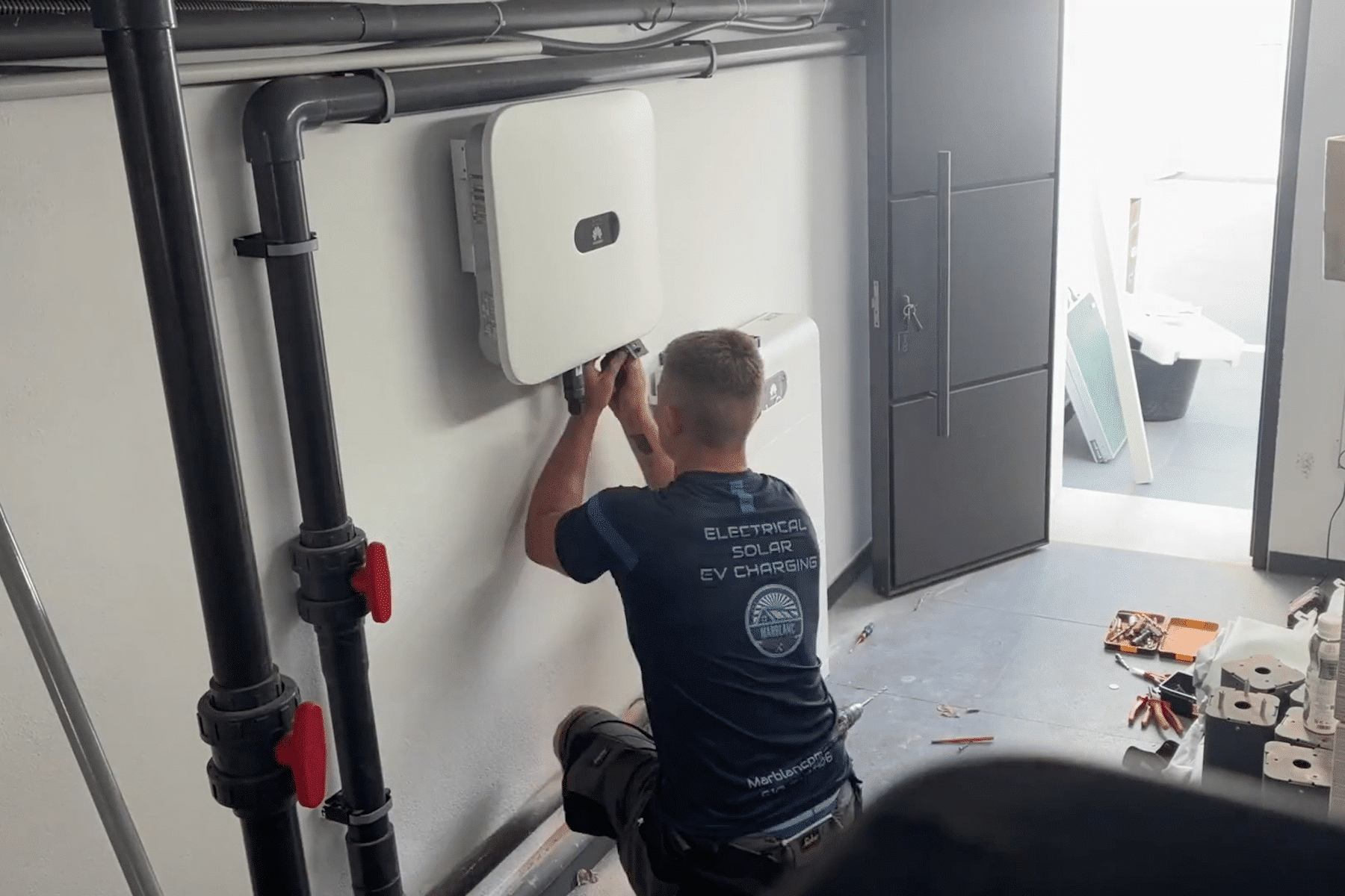 Un hombre agachado, trabajando o inspeccionando un aparato blanco montado en la pared con tubos alrededor en una habitación.