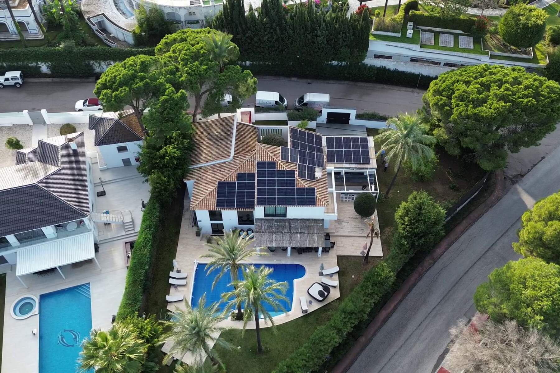 Aerial view of a villa with many solar panels on the roof, surrounding greenery, and a rectangular blue swimming pool in the yard.